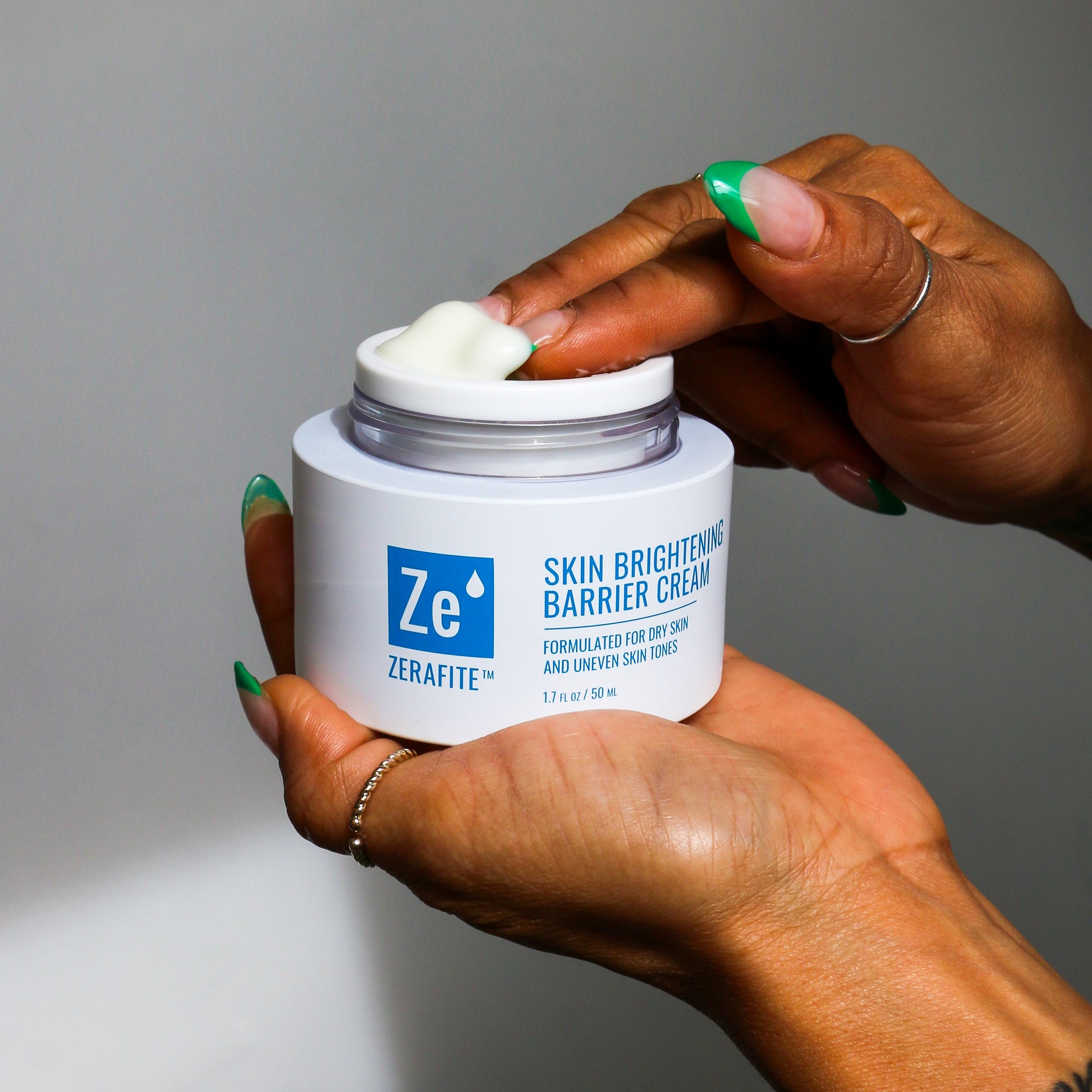 A person's hands holding a white jar of Zerafite Skin Brightening Barrier Cream, with a dollop of the white cream visible on the lid, against a neutral gray background.