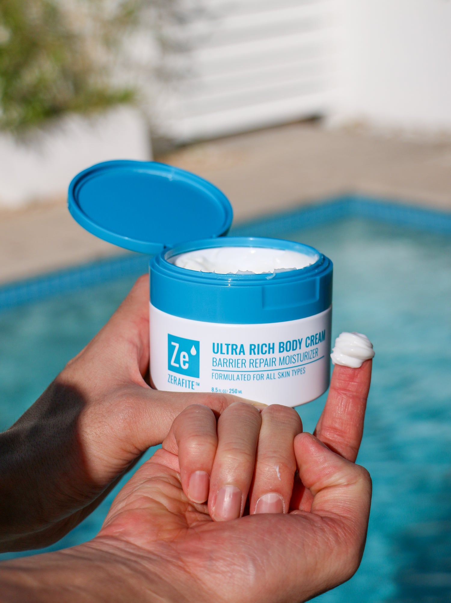 Person holding an open jar of Zerafite Ultra Rich Body Cream by a pool with the product on their finger tip to show texture